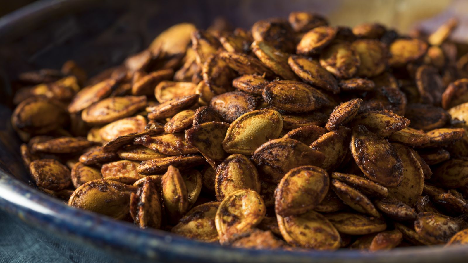 A close-up and focused shot of roasted and spiced squash seeds in a dark colored container placed on top of a wooden surface indoors